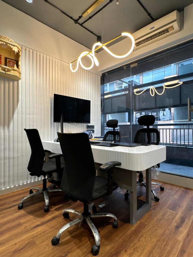A modern Ahmedabad office space featuring a white desk with multiple black office chairs, a large mounted monitor, and unique curved overhead lighting, beside a window with blinds