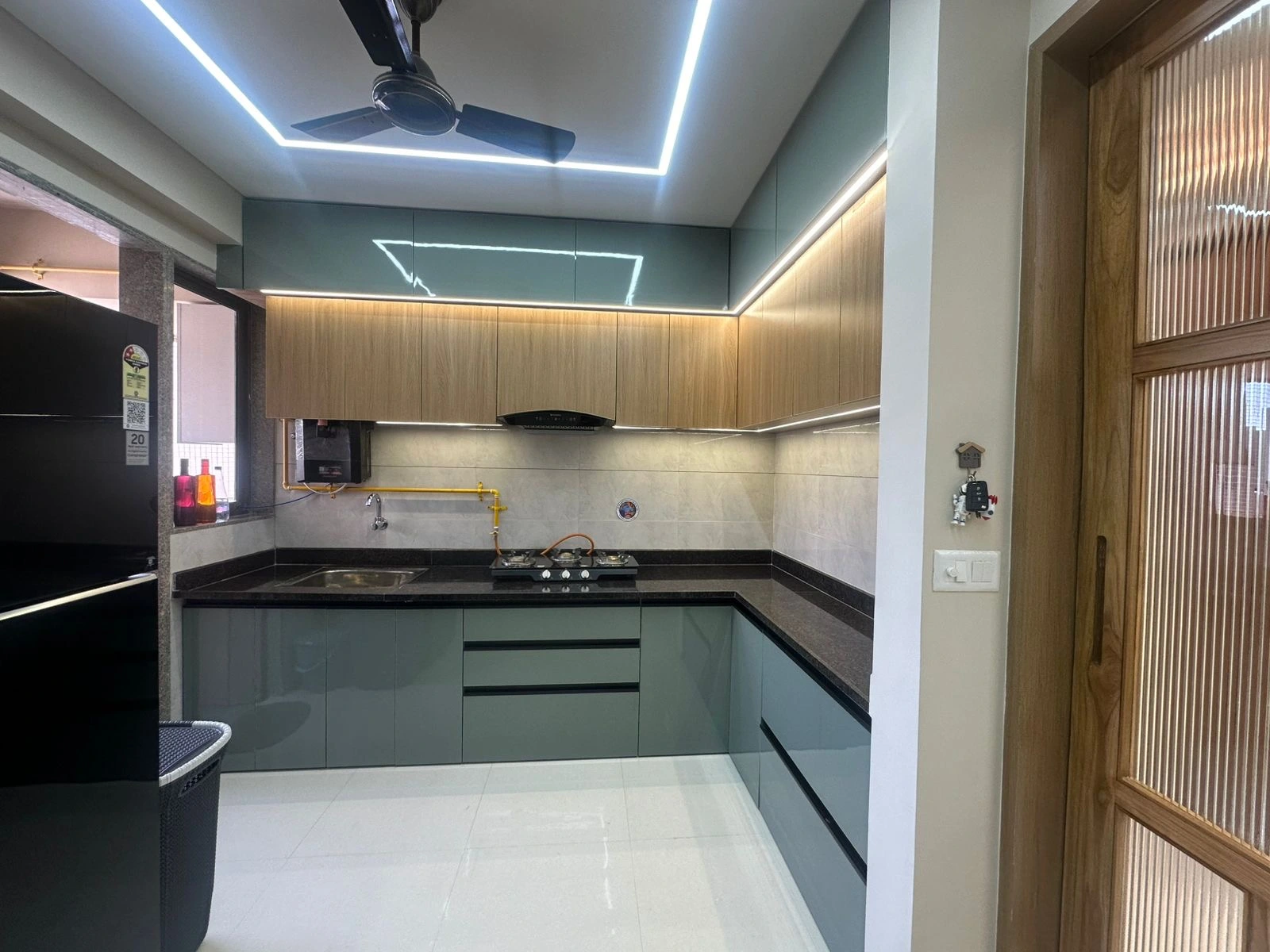 Modern L-shaped modular kitchen design with pastel green cabinets and wood upper storage by J Designs in Ahmedabad